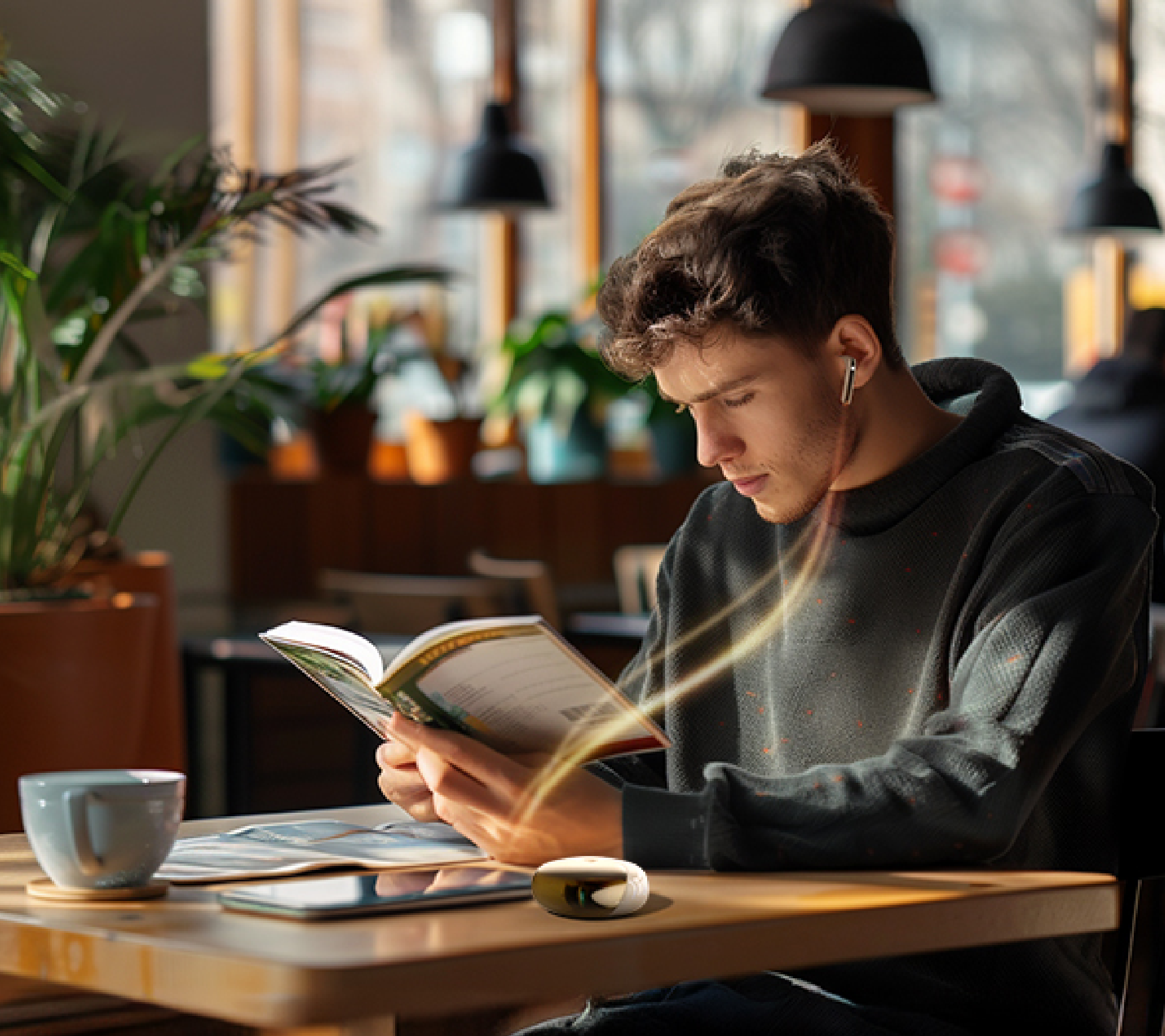 Jovem lendo livro em cafeteria com fones sem fio e xícara de café na mesa, ambiente aconchegante e iluminado.