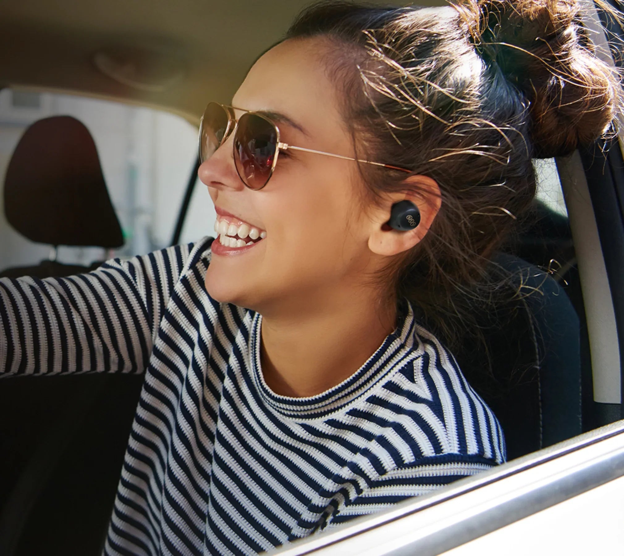 Mulher sorrindo no carro usando fones de ouvido sem fio e óculos escuros, aproveitando música ou chamada em movimento.