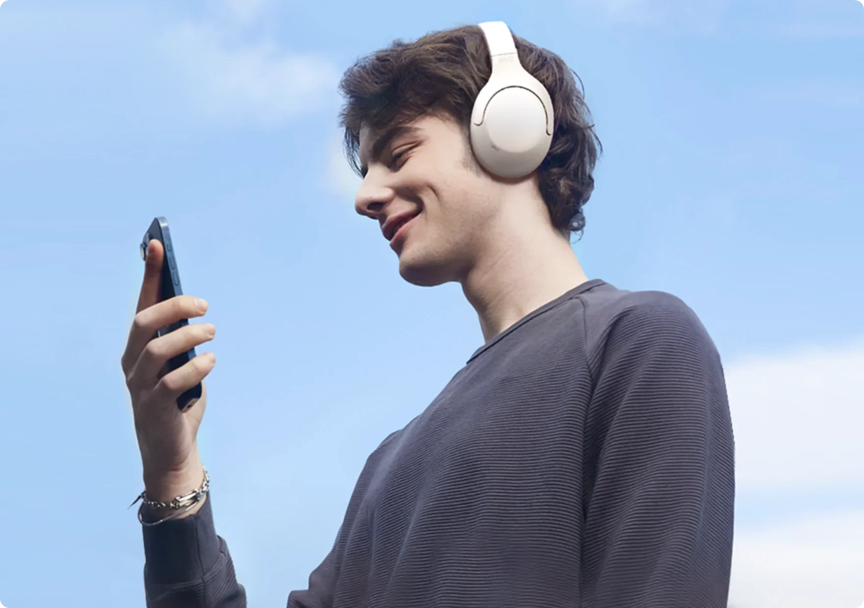 Jovem sorridente usando fones de ouvido brancos e segurando smartphone, aproveitando música ao ar livre sob céu azul.