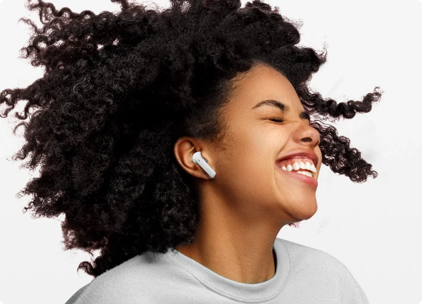 Mulher sorrindo com fones de ouvido sem fio brancos, cabelos cacheados ao vento, fundo branco. Produto tecnológico para músic