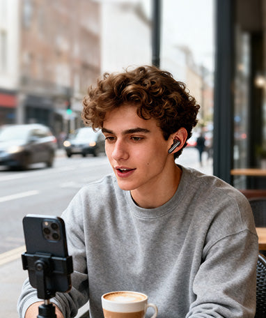 Jovem usando fones de ouvido sem fio em café, gravando vídeo com smartphone. Tecnologia moderna para conteúdo criativo e comu