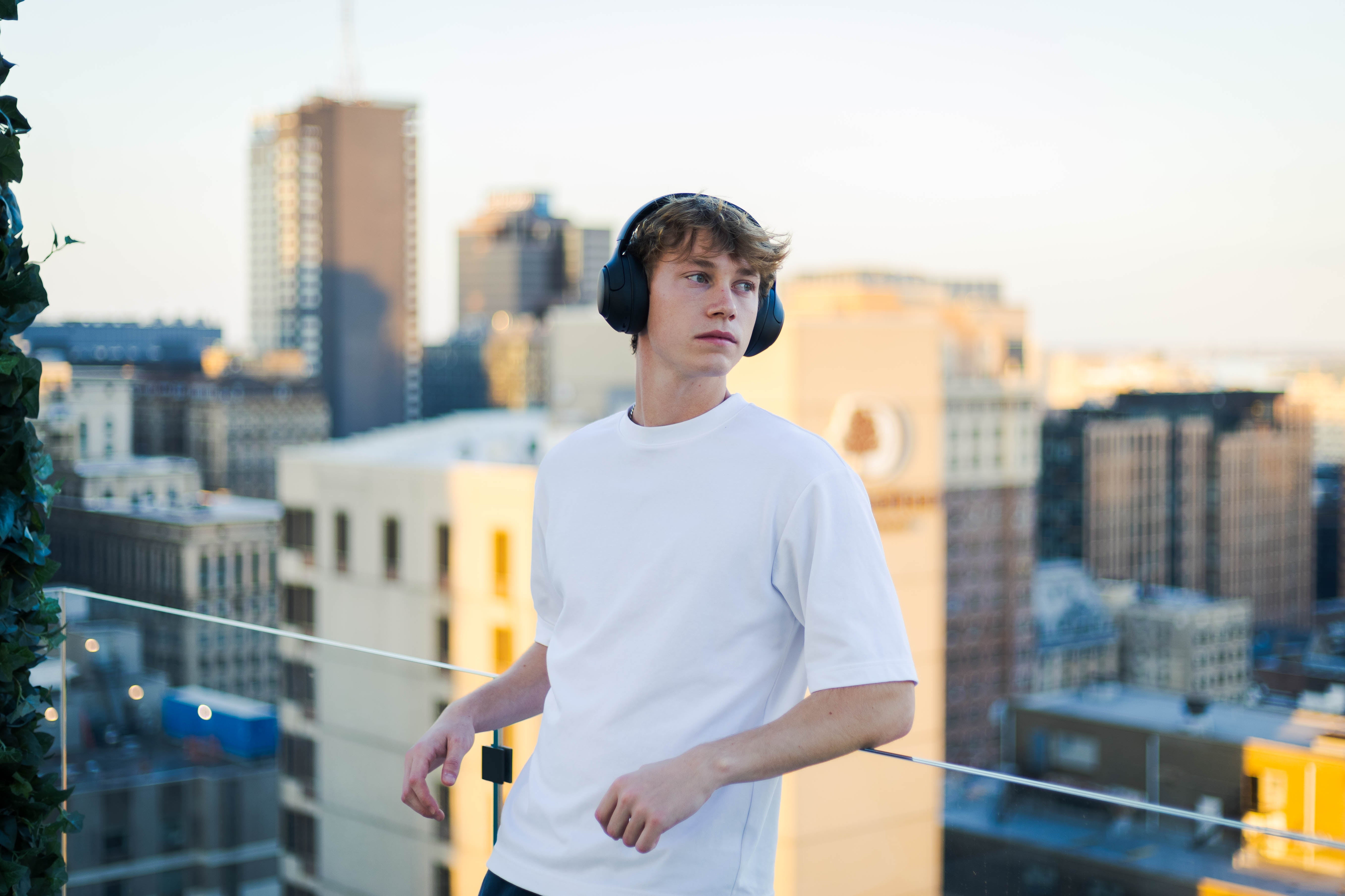 Jovem usa fones de ouvido sem fio em rooftop com vista da cidade ao pôr do sol.